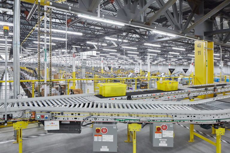 An expansive warehouse in Troutdale, OR, features a complex system of conveyor belts and is brightly lit with overhead fluorescent lights. For expert electrician services in the nearby area of Vancouver, WA, we specialize in handling such industrial electrical systems efficiently.