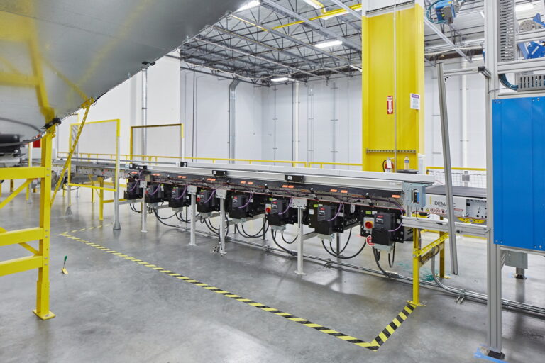 A modern industrial distribution center in Troutdale features advanced electronic machinery and equipment along a conveyor belt. With its yellow safety barriers, metal beams, and overhead lighting, the facility prioritizes efficiency and safety. For professional electrician services to maintain or upgrade such systems in the Vancouver, WA area, expertise is essential to ensure seamless operations.