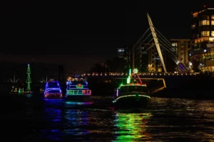 Boats with expertly installed Christmas lights travel a Vancouver, WA river at night; bridge and buildings also feature professional lighting.