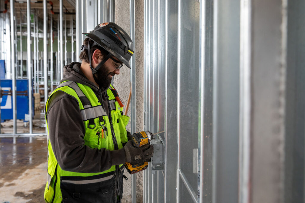 An electrician in Vancouver, WA, wearing a green safety vest and black helmet, skillfully operates a power drill on a metal wall frame amidst partially constructed walls and building materials.