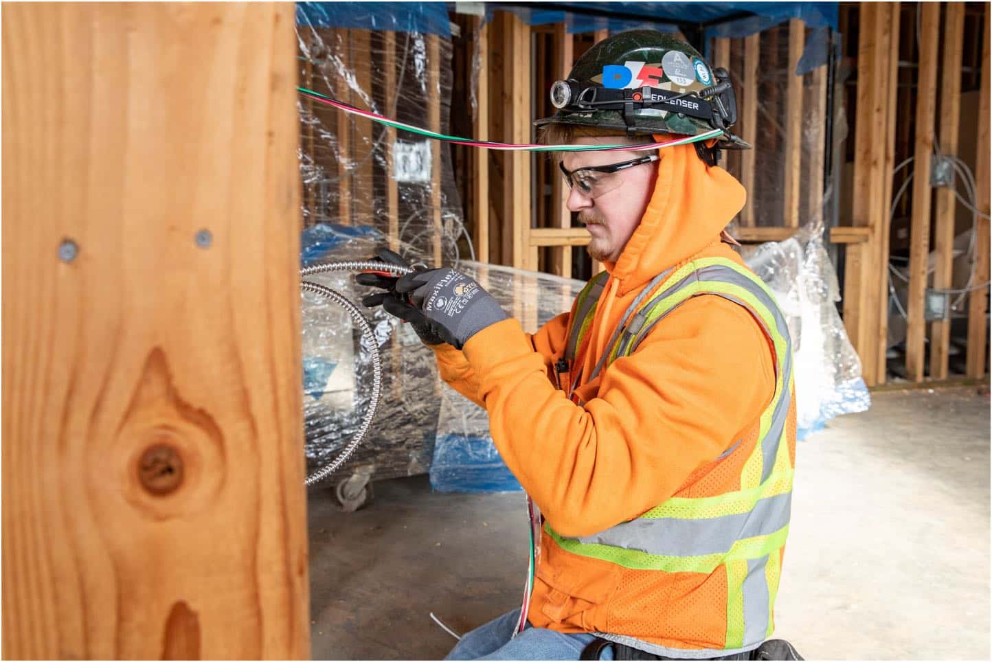 An electrician wearing safety gear, including a hard hat and safety glasses, is installing electrical wiring in a wooden framework in Vancouver, WA. The background features unfinished construction with wooden beams.