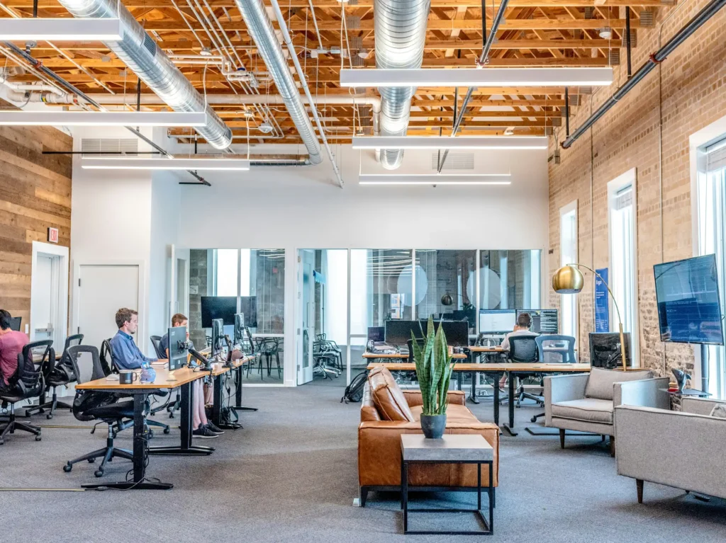 An open office space with people working at desks equipped with computers. A seating area with a couch and a plant is in the foreground. High exposed ceilings, installed by a Vancouver electrician, feature industrial fixtures overhead.