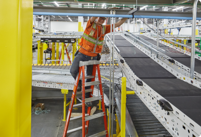 Prairie Electric technician on a red ladder completes an electrical installation project in an industrial setting with conveyor belts and yellow structures in the background.