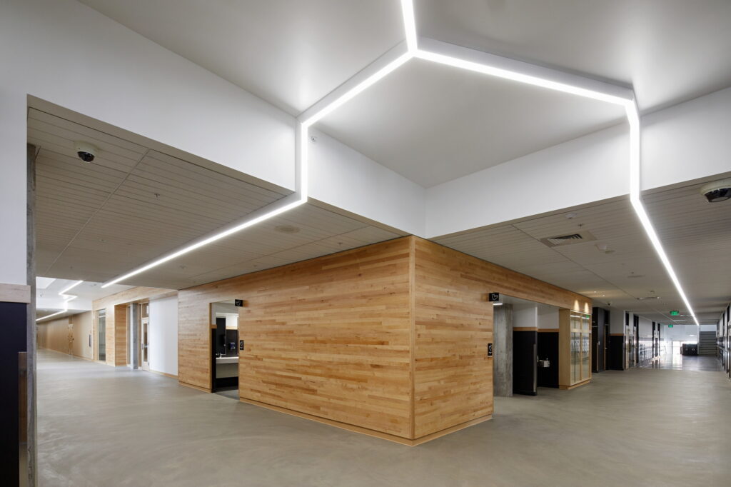 Grant High School in Portland, OR boasts a modern interior with light wooden walls and polished concrete floors. The strategically placed LED lighting creates geometric patterns on the ceiling, enhancing visibility and guiding students through the hallways, emphasizing the importance of efficient electrical design in educational settings.