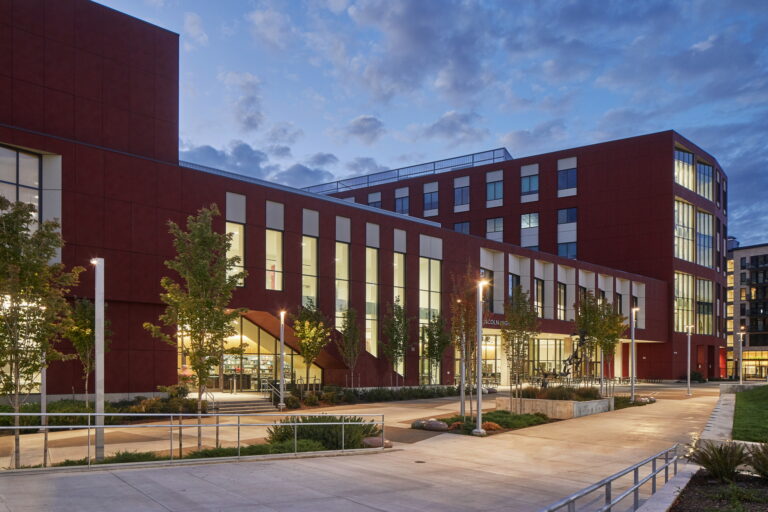 Lincoln High School in Portland, OR showcases a modern architectural design with large windows and red exterior walls. The building is equipped with professional electrician services ensuring effective lighting throughout the interior and exterior, creating a safe and welcoming environment under the partly cloudy sky at dusk.