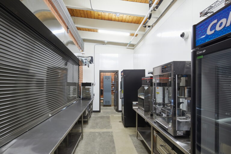 The commercial kitchen at Lincoln High School features stainless steel appliances and fixtures, with beverage machines and equipment lining one side. The clean, well-lit space reflects an organized industrial atmosphere ideal for efficient electrical work.