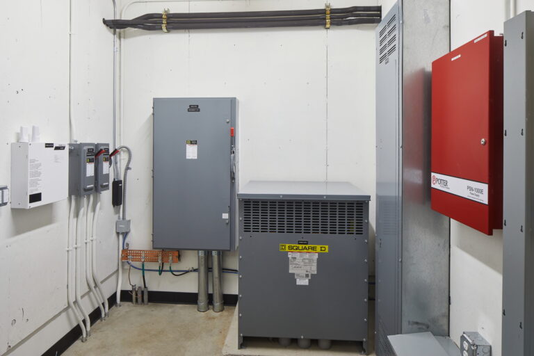 The utility room at Lincoln High School contains essential electrical equipment such as a grey "Square D" box and wall-mounted panels crucial for operations. It includes assorted wiring and a red panel labeled "Protective Power Systems and Controls.