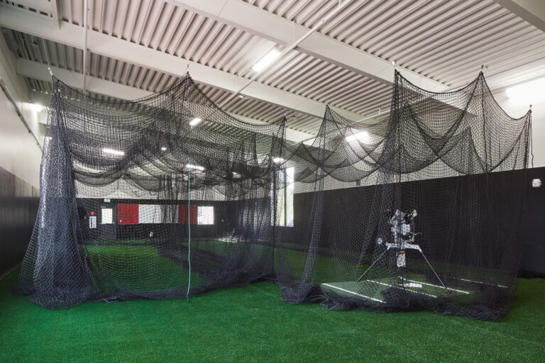 A large indoor batting cage at Lincoln High School features black netting and an artificial turf floor. A centrally positioned pitching machine and natural light streaming through a side window highlight the need for expert electrician services to ensure proper lighting installation and maintenance.