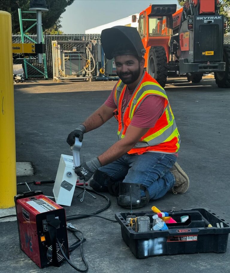 A worker wearing a safety vest and helmet kneels on the ground, handling equipment to help prevent power outages. A toolbox is nearby. Heavy machinery and fencing are visible in the background.