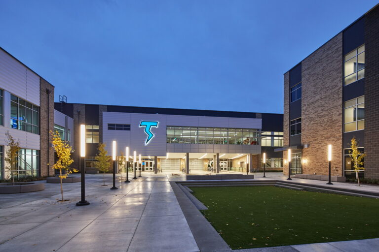 A modern high school exterior in Vancouver, WA, featuring an illuminated "T" sign, exemplifies sophisticated electrical work. This architectural blend of glass, brick, and metal is enhanced by strategic lighting throughout the courtyard with bollards and accentuated paths against a mountain backdrop.