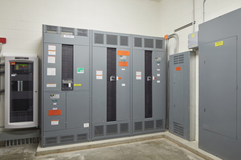 The image depicts a room at Mountain View High School in Vancouver, WA, highlighting gray electrical panels on white walls. These panels, equipped with various switches and labels, are characteristic of the electrician services required for their installation and maintenance. A smaller control box is also visible alongside cables running across the ceiling and walls.