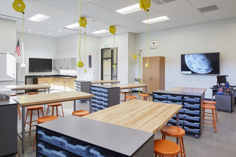A modern classroom with workstations, orange stools, and overhead retractable power cords. There is a television displaying a moon image on the right wall and cabinets in the background.