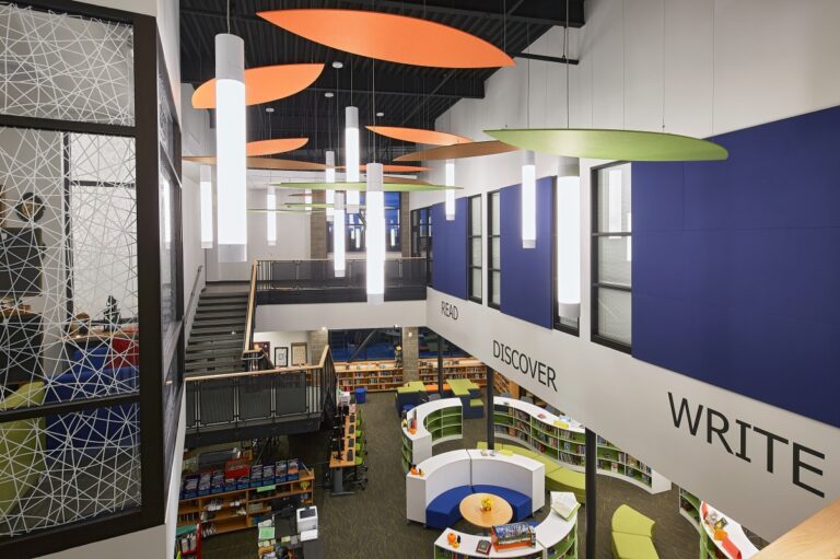 A modern library interior featuring colorful overhead panels, a central circular reading area, and bookshelves lining the lower level. Large windows and a staircase are visible on the left side.