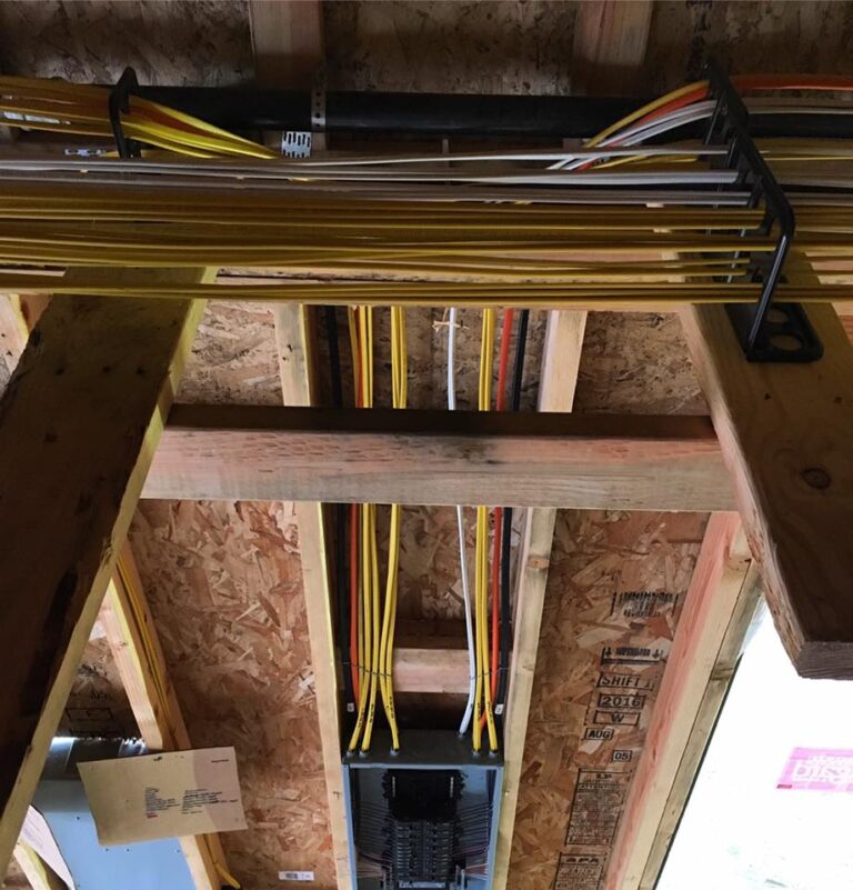 In Vancouver, WA, yellow and white electrical wires are neatly secured with black brackets along wooden framing and enter an electrical panel on an unfinished plywood wall. This organized installation by professional electricians helps prevent common electrical issues in construction settings.