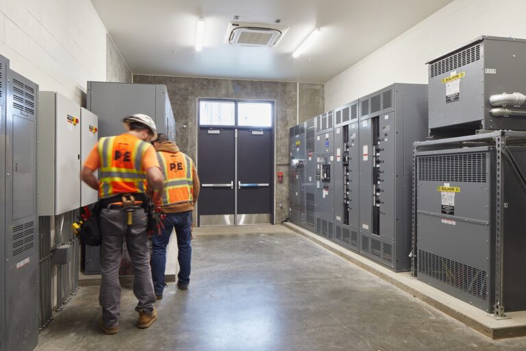 Two workers in reflective vests and hard hats stand in a room with large electrical panels.