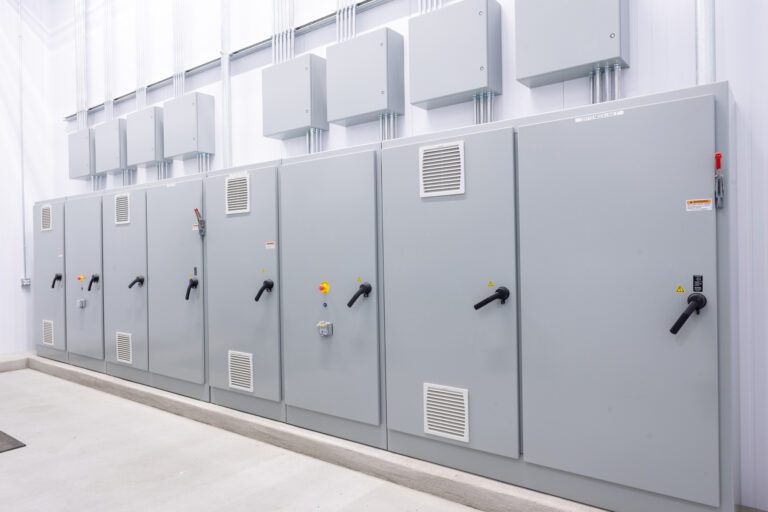 A row of large gray electrical panels with handles and vents is neatly arranged at Reser Fine Foods. These panels, equipped with warning labels and smaller boxes above, highlight the need for professional electrician services to ensure safety and efficiency.