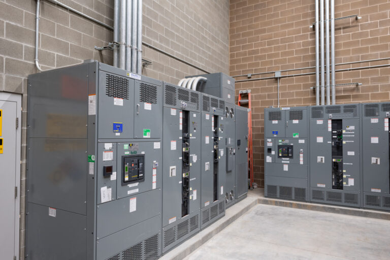 The image shows an industrial electrical room with large gray panels equipped with switches and meters. Metal conduits run from the panels to the ceiling against beige and gray concrete block walls.