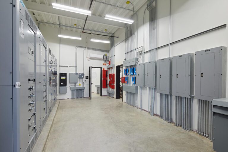 A clean, well-lit industrial control room featuring electrical panels, conduit, and various control units mounted on the walls and cabinets. The floor is bare concrete, and the ceiling is made of white panels.