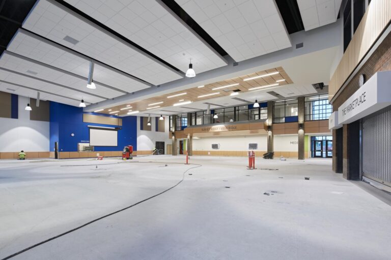 A large, empty, modern indoor space with high ceilings, lights, and under-construction areas. "Mercyhealth" signage is visible above a balcony and a space labeled "Main Entrance.
