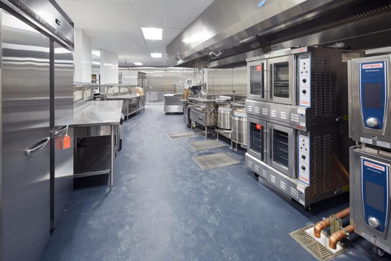 Commercial kitchen with stainless steel appliances, including ovens, refrigerators, and work tables. The room is clean and spacious with a blue epoxy resin floor and overhead exhaust hoods.