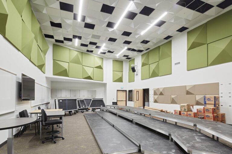 A modern classroom with modular acoustic panels on the walls, fluorescent lighting on the ceiling, several chairs and desks, a television screen, and a storage area with boxes and equipment.