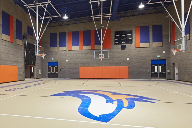 An indoor basketball court with a central logo on the floor, multiple hoops, scoreboards, and padded walls; surrounded by colorful panels and doors on the sides.