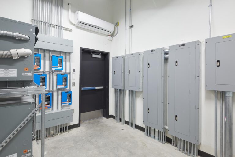 A room with multiple electrical panels and conduit pipes mounted on the walls, an HVAC unit on the ceiling, and a closed black door.