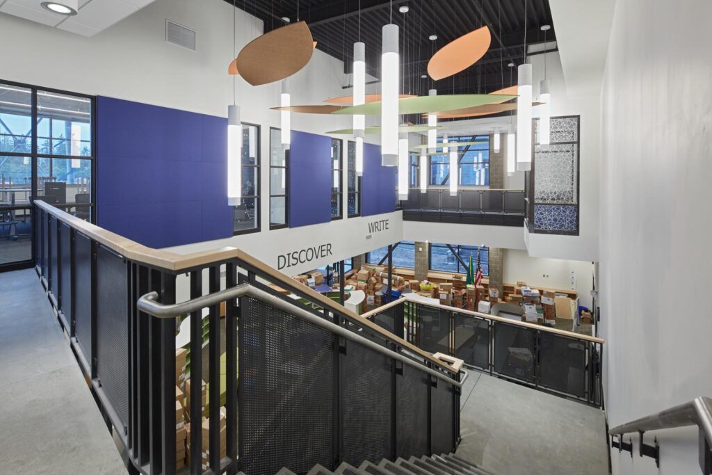 Interior view of a modern, multi-level library with stairs leading down to a main area. Bookshelves, study areas, and large windows are visible, along with words "DISCOVER" and "WRITE" on a wall.