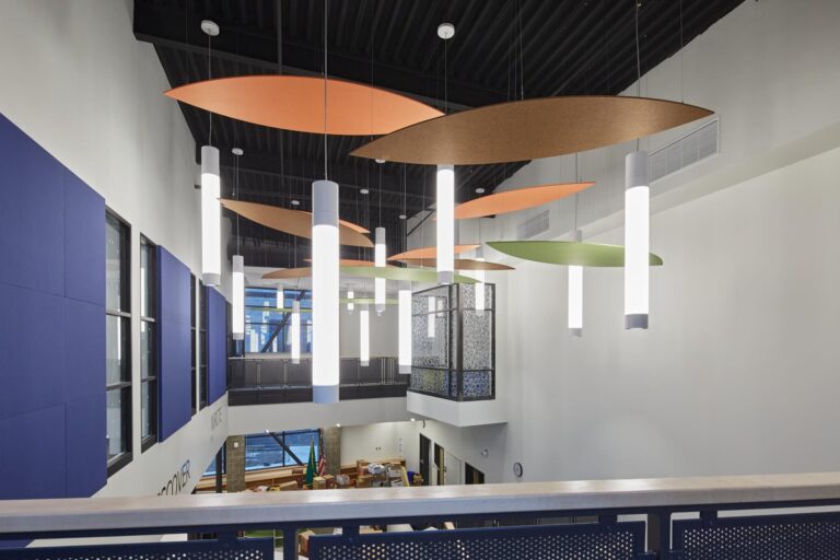A modern interior space with colorful leaf-shaped ceiling panels, cylindrical hanging lights, large windows, and a view of furniture and decor below.