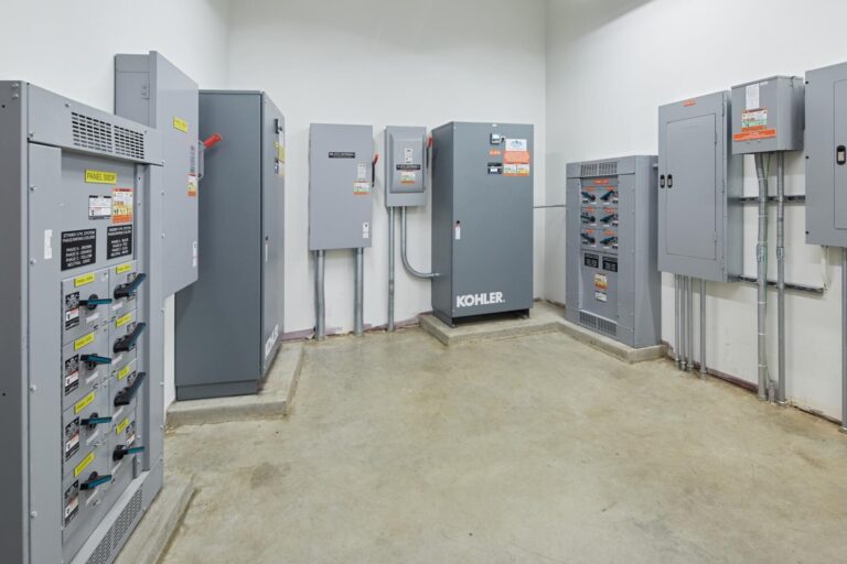 A room with electrical panels and equipment, including units labeled Kohler, against white walls with a concrete floor. Labels and switches are visible on the various panels.