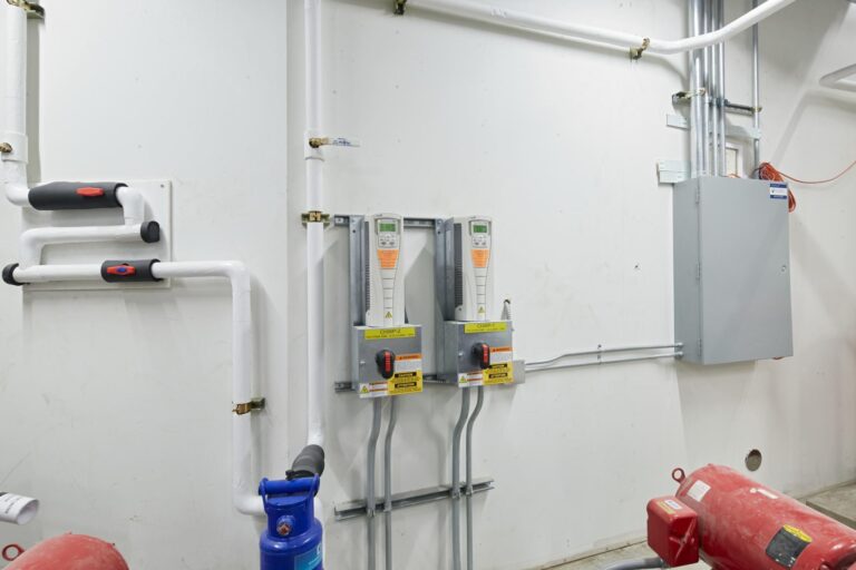 A wall-mounted control panel in a utilities room, featuring two digital meters, various pipes, valves, an electrical fuse box, and other plumbing components.