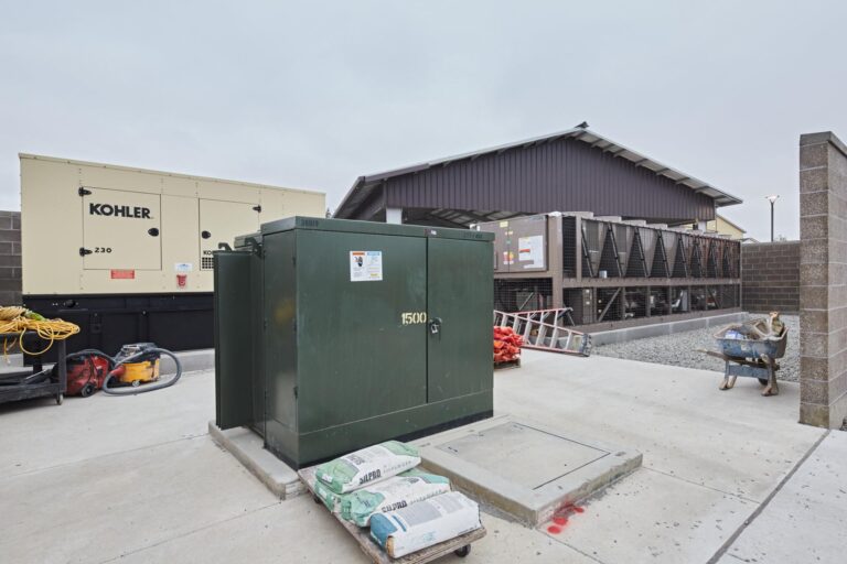 Outdoor utility area with industrial generators, electrical box, building materials, and equipment around a concrete pad in front of a large, dark brown structure.