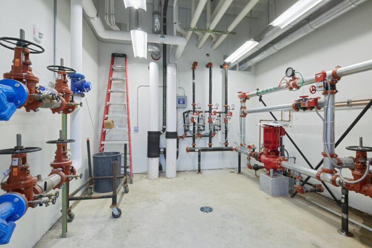 A utility room with various pipes, valves, and mechanical equipment, a red ladder leaning against the wall, and fluorescent lighting overhead.