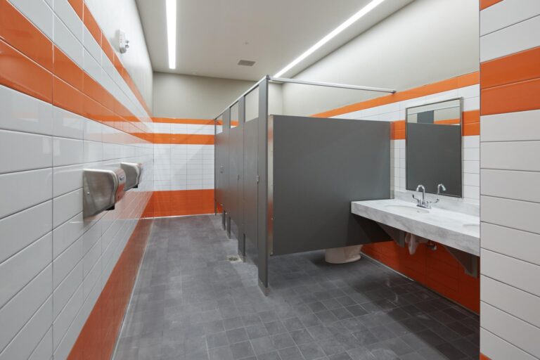 A public restroom with gray stall doors, white and orange tiled walls, a wall-mounted mirror, a sink with two faucets, and two stainless steel hand dryers on the left.