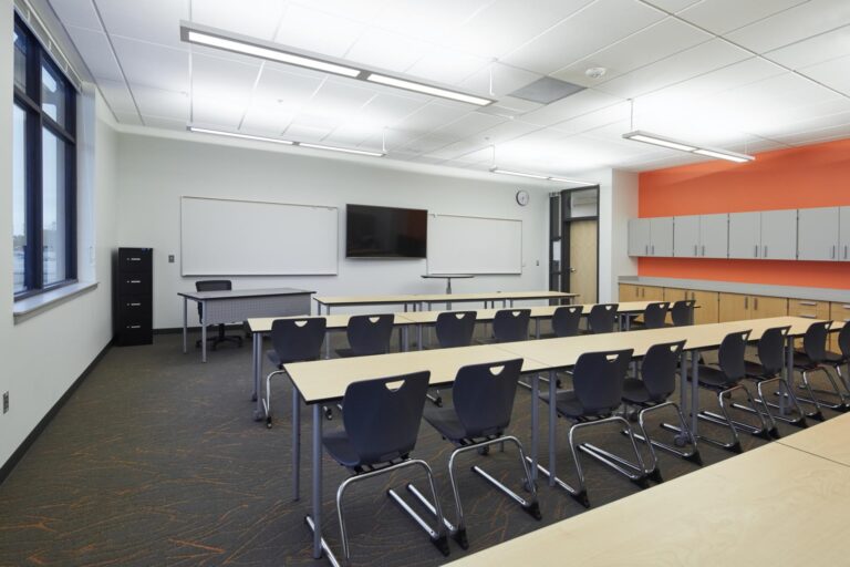 A modern classroom with empty desks and chairs, a large window, whiteboards, a wall-mounted TV, and storage cabinets along an orange wall.