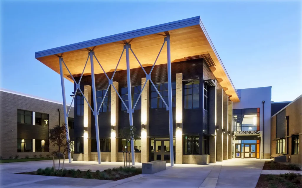 A modern two-story building with large windows and illuminated pillars at dusk, featuring a wood and metal awning. The sign above the entrance reads "Commons." The stunning lighting design was expertly crafted by a Vancouver electrician.