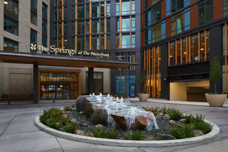 Round fountain with cascading water in front of a modern building entrance labeled "The Springs at The Waterfront." Greenery surrounds the fountain.