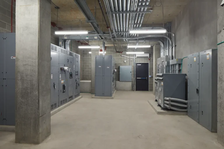 An industrial room with concrete walls, electrical panels, conduit pipes on the ceiling, and a metal door. Bright lighting illuminates the space.