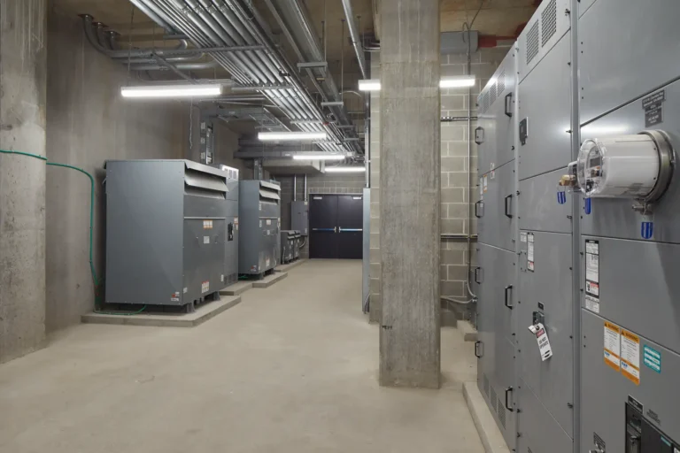 Industrial room with large metal equipment and control panels, concrete walls, and overhead pipes. Fluorescent lights illuminate the space.