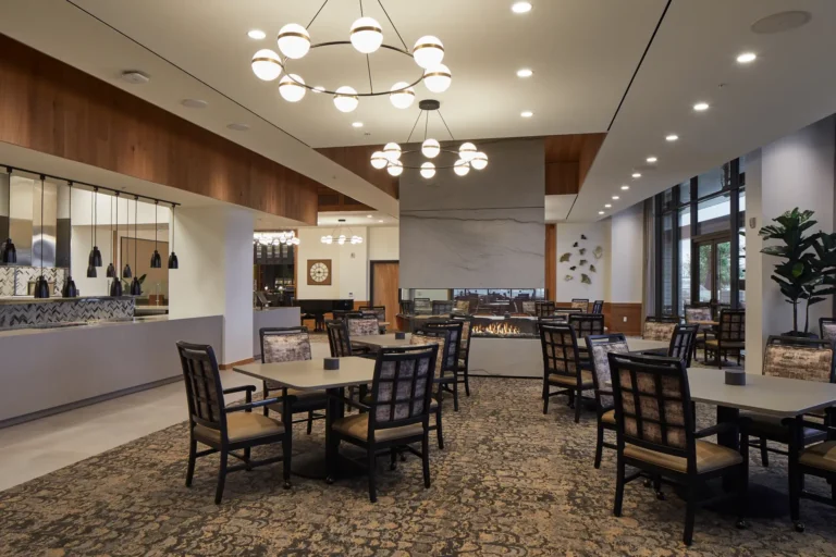 A modern dining area with rectangular tables and padded chairs, overhead lighting fixtures, and a fireplace. Large windows in the background let in natural light.