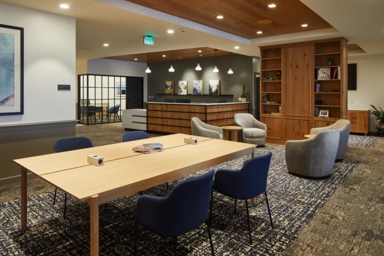 Modern office lounge with a wooden table, blue chairs, a seating area with gray armchairs, bookshelves, and overhead lighting. Walls feature framed artwork and a partition with glass panels.