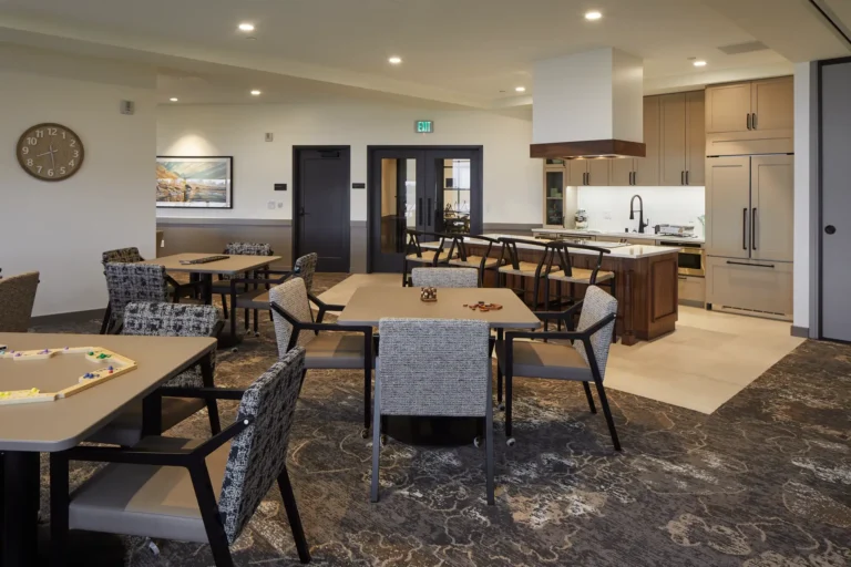A modern dining area with several tables and chairs, a kitchen island with stools in the background, and decorative lighting in an open, well-lit space.