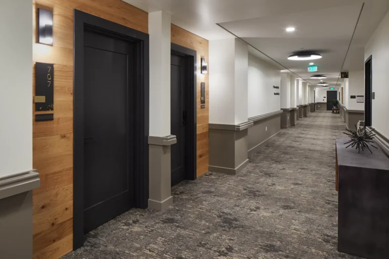 A modern hotel hallway with closed dark doors and wooden accents. Neutral-toned carpet runs the length of the corridor, and wall-mounted lights illuminate the space.