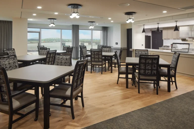 A modern dining area with several tables and chairs, featuring wooden flooring and overhead lights, and a window view of a landscape.