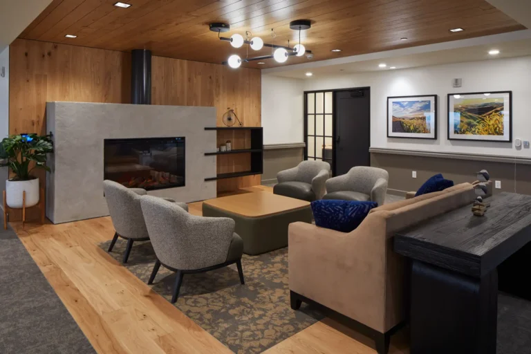 Modern living room with a fireplace, surrounded by a beige and blue sofa, two grey chairs, a coffee table, wall art, and overhead lighting.