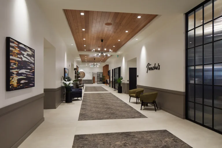 Modern hallway with wooden ceiling, chandelier, abstract art, and "Goodrich’s" on the wall. Two green chairs and potted plants line the space.