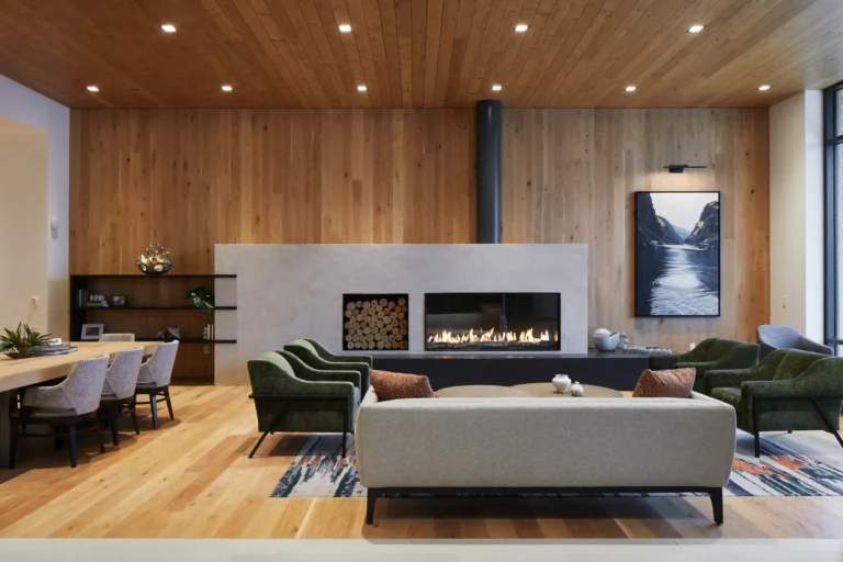 Modern living room with wooden ceiling and floor, a fireplace, two sofas, four armchairs, and a rug. Large windows on the right and artwork on the wall.
