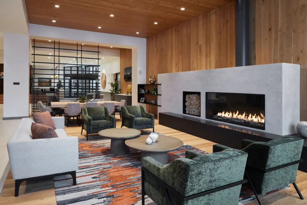 Modern living room with a large fireplace, green and gray furniture, wooden walls, and a geometric rug. Ceiling lights illuminate the space.