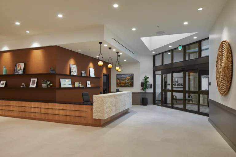 Modern hotel lobby with a reception desk, decorative shelves, hanging lights, and glass entrance doors.