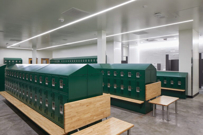 The image showcases the well-lit locker room at Tigard High School highlighting rows of dark green lockers with wooden benches. The clean and organized space might benefit from an electrician's services for optimal lighting installation and maintenance.